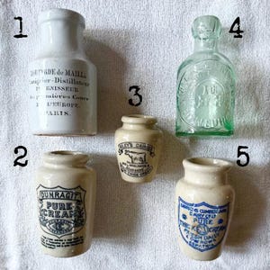 May include: An assortment of antique bottles and jars, including a white ceramic bottle with French text, a green glass bottle, and several small cream jars with labels. The items are arranged on a white surface.