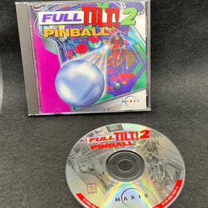 May include: A Full Tilt! 2 Pinball PC game in a clear plastic case. The game's cover art features a pinball machine with a large blue ball and a figure in a red suit. The CD is also visible.