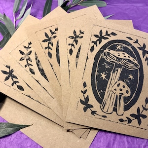 Puede incluir: Un conjunto de seis tarjetas de papel kraft marrón con ilustraciones de tinta negra de setas y hojas. Las tarjetas están apiladas en forma de abanico.