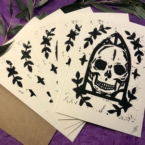 Peut inclure: Ensemble de cinq cartes d'impression noir et blanc représentant un crâne avec des accents floraux. Le crâne porte une capuche et est entouré de feuilles et d'étoiles.