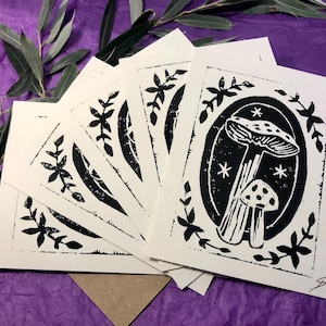 Puede incluir: Un juego de cinco tarjetas de felicitación en blanco con ilustraciones en tinta negra de hongos y follaje. Las tarjetas están impresas en cartulina blanca y tienen una superficie ligeramente texturizada.