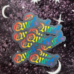 Puede incluir: Primer plano de varias pegatinas holográficas arcoíris con la palabra "Queer" en una fuente gótica.