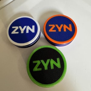 以下が含まれることがあります： 「ZYN」の文字がプリントされた、3つの丸いカラフルなオブジェクト。1つは白文字の青色、もう1つはオレンジ文字の青色、3つ目は緑文字の黒色です。白い表面に置かれています。