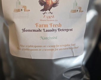 Op de afbeelding: Een zilveren zak met zelfgemaakt wasmiddel. Het etiket toont een vogelillustratie en de tekst "Farm Fresh Homemade Laundry Detergent Narcissist". Instructies suggereren het gebruik van 2 eetlepels of 1 schep voor een normale lading.