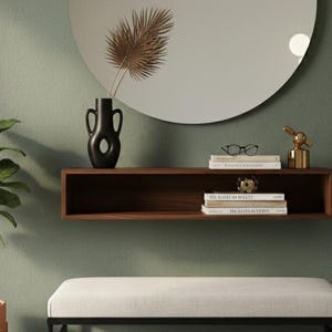 Op de afbeelding: Een modern interieur met een grote ronde spiegel, een zwevend houten rek met decoratie en een bank. Een potplant en een zwarte vaas met gedroogde bladeren completeren het geheel. De muren zijn groen.
