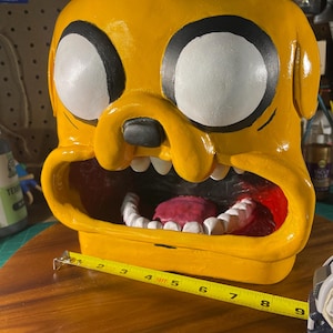 Op de afbeelding: Een gele sculptuur van een cartoonkarakterkop met grote witte ogen omlijnd met zwart. De open mond onthult witte tanden en een rode tong. De sculptuur is ongeveer 23 cm breed.