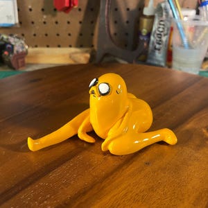 May include: A handcrafted, yellow Jake the Dog figurine from Adventure Time. The figure is posed in a lying down position with his arms and legs extended. The figure has black and white eyes and a small smile.