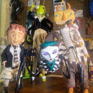 Könnte beinhalten: Eine Gruppe skurriler, handgefertigter Kunstpuppen. Die Puppen umfassen einen Jungen in einem Blazer, eine Heuschrecke im Anzug, eine spinnenartige Figur und einen käferartigen Charakter. Die Puppen sind auf einer Holzoberfläche vor einem Hintergrund aus Lochplatten aufgestellt.
