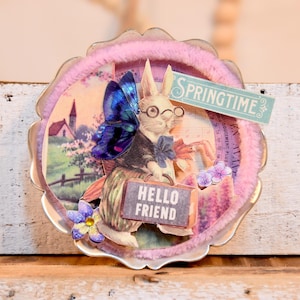 Op de afbeelding: Een ronde, roze en witte, decoratieve metalen versiering met een pluche roze rand. De versiering heeft een papieren uitsnijding van een konijn met een bril en een blauwe vlinder op zijn rug. Het konijn houdt een koffer vast waarop "Hello Friend" staat. De versiering heeft ook een papieren uitsnijding van een kerk en een bord met de tekst "Springtime".