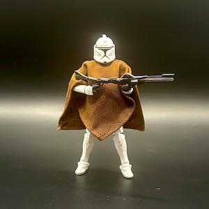 May include: A white plastic action figure of a soldier wearing a brown fabric poncho over white armor. The figure has a white helmet with black visor details and is holding a long black blaster rifle in a firing position with both hands.