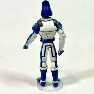 May include: A standing action figure viewed from the back, featuring a blue and white armored suit with a tall, rounded blue helmet. The figure has white shoulder plates, blue arm details, and stands on a clear circular base against a white background.