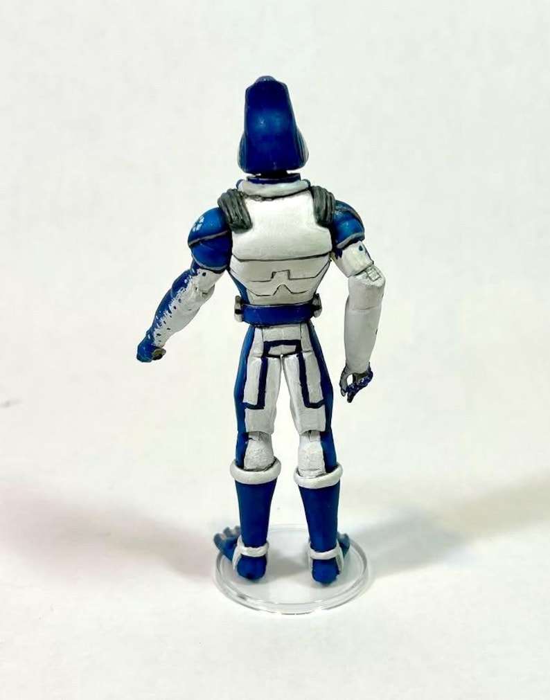 May include: A standing action figure viewed from the back, featuring a blue and white armored suit with a tall, rounded blue helmet. The figure has white shoulder plates, blue arm details, and stands on a clear circular base against a white background.