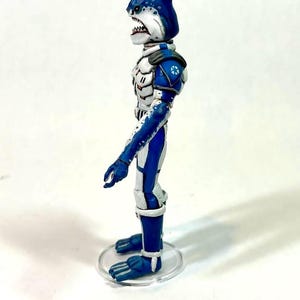 May include: A side profile of a humanoid shark action figure with blue and white skin, wearing white and blue armor with orange accents. The figure has a dorsal fin on its head, sharp teeth, and stands on a clear circular plastic base.