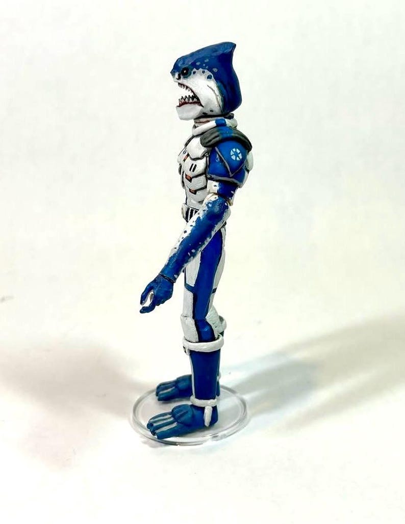 May include: A side profile of a humanoid shark action figure with blue and white skin, wearing white and blue armor with orange accents. The figure has a dorsal fin on its head, sharp teeth, and stands on a clear circular plastic base.