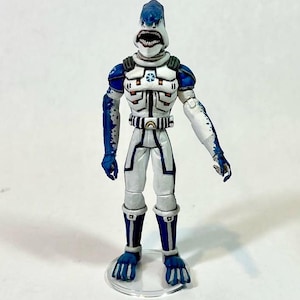 May include: A humanoid shark action figure wearing a white and blue armored suit with a high collar, chest emblem, and belt. The figure features a shark head with an open mouth showing teeth, blue limbs with white accents, and stands on a clear base.