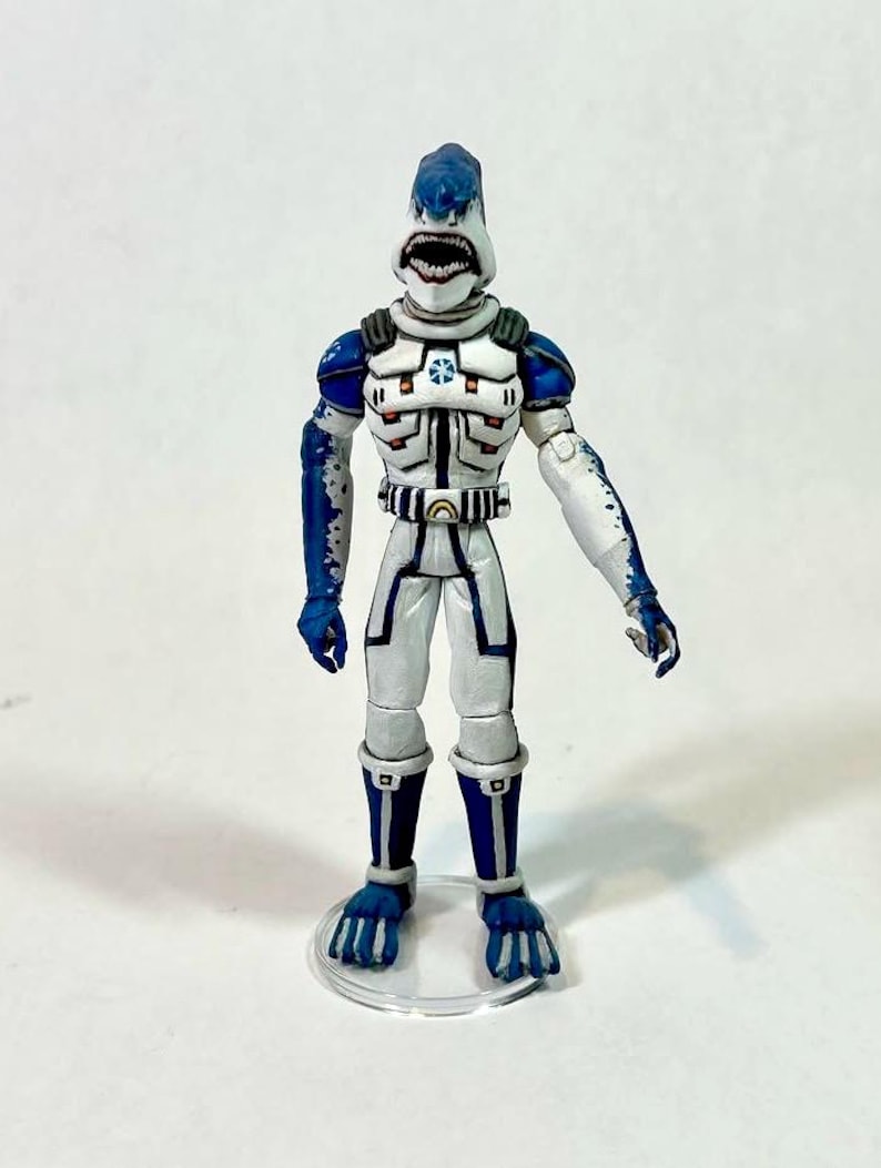 May include: A humanoid shark action figure wearing a white and blue armored suit with a high collar, chest emblem, and belt. The figure features a shark head with an open mouth showing teeth, blue limbs with white accents, and stands on a clear base.
