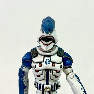 May include: A humanoid action figure with a shark head featuring a blue top and white lower jaw with visible teeth. The figure wears white and blue armor with orange accents, a blue star emblem on the chest, and a belt with a yellow circular buckle.