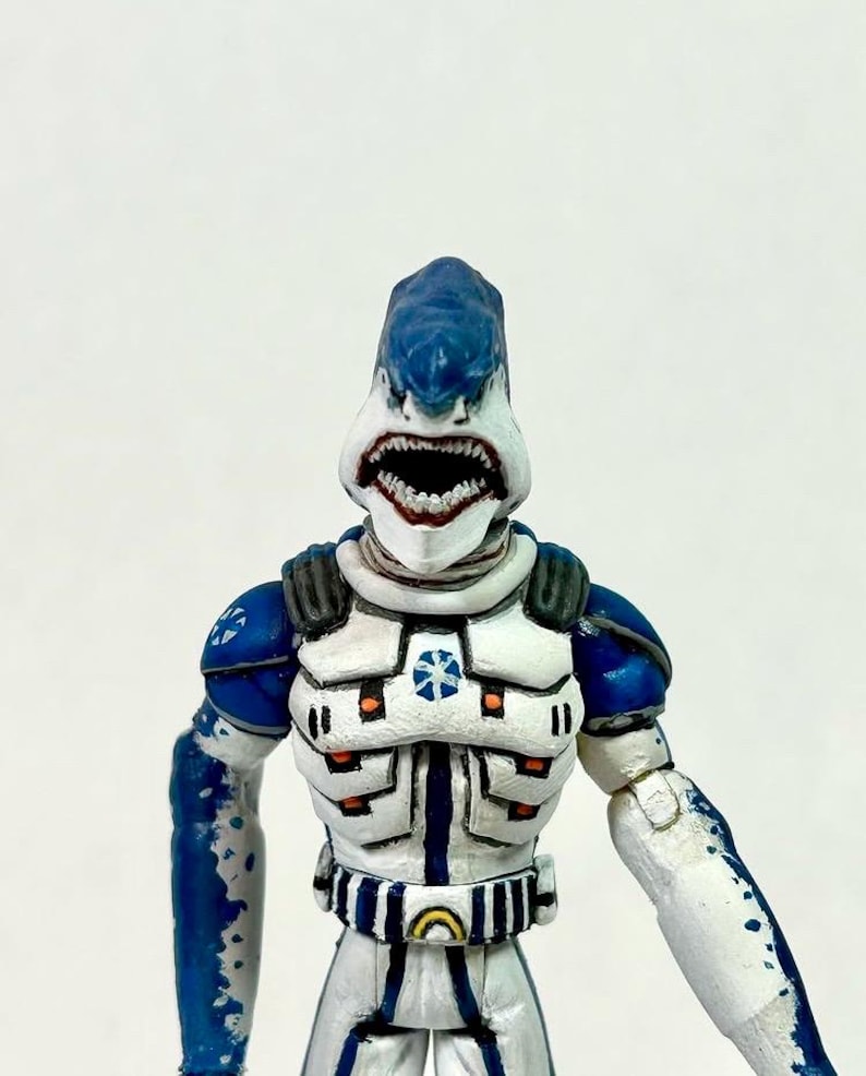 May include: A humanoid action figure with a shark head featuring a blue top and white lower jaw with visible teeth. The figure wears white and blue armor with orange accents, a blue star emblem on the chest, and a belt with a yellow circular buckle.