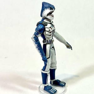 May include: A standing action figure of a shark-headed humanoid character wearing a white and blue armored suit. The figure features a blue shark head with visible teeth, blue and white patterned limbs, and a white chest plate with blue accents.