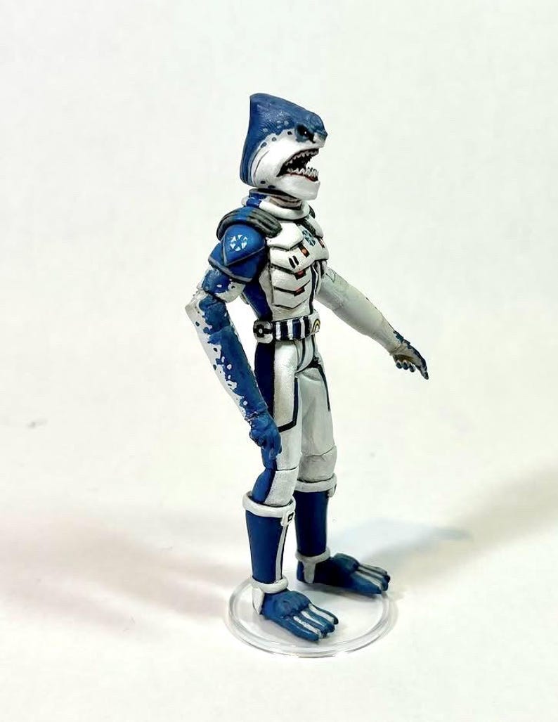 May include: A standing action figure of a shark-headed humanoid character wearing a white and blue armored suit. The figure features a blue shark head with visible teeth, blue and white patterned limbs, and a white chest plate with blue accents.