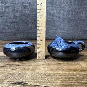 Puede incluir: Dos pequeñas piezas de cerámica azul oscuro y negro. Una es un cuenco redondo, la otra es una jarrita con asa. Una regla de madera está al lado de las piezas, midiendo aproximadamente 10 cm.