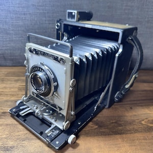 May include: A vintage black and silver Speed Graphic camera with a bellows design. The camera has a lens with the text "Graflex" and "135mm" on it. The camera is on a wooden surface.