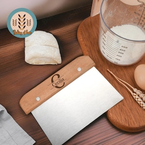 May include: A wooden and stainless steel dough cutter with a personalized inscription that reads "Cecilia" on the handle. The cutter is on a wooden cutting board with a ball of dough, a measuring cup of flour, and an egg.