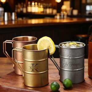 May include: Three metal barrel-shaped mugs with handles, featuring a copper, gold, and silver finish. The gold mug is engraved with the letter K and the name Kathryne, while the silver mug features the text La Frasquita and contains ice and a lemon slice.
