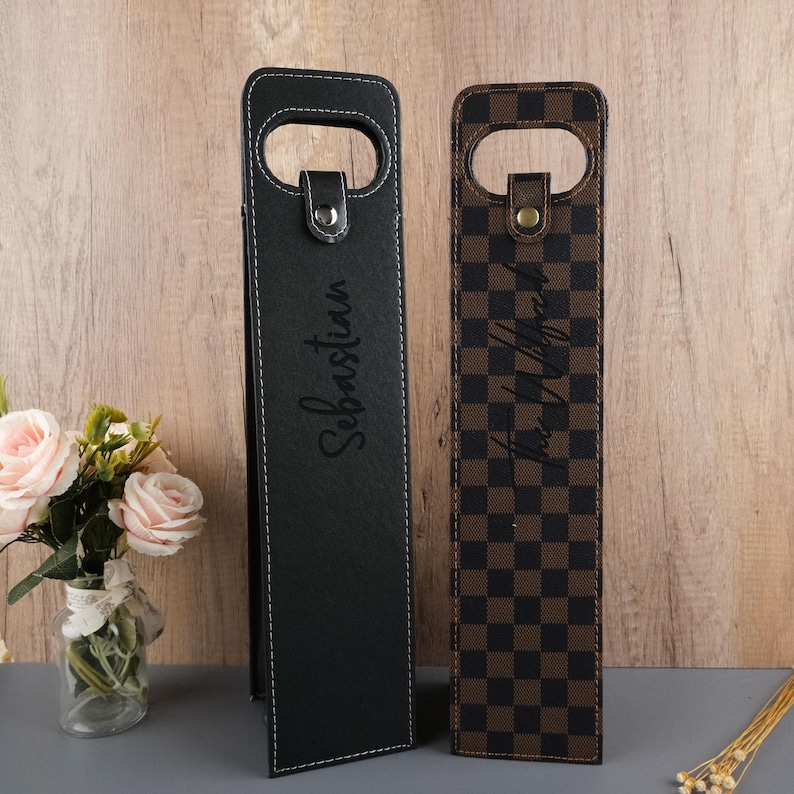 K&ouml;nnte beinhalten: Zwei schwarze Leder-Weinflaschentaschen mit personalisierten Namen. Die Tasche links hat den Namen "Sebastian" eingraviert. Die Tasche rechts hat ein braunes und schwarzes Karomuster und den Namen "Tina Whitehead" eingraviert.
