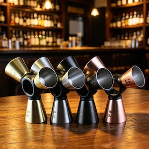 May include: Four multi-cup cocktail jiggers in gold, silver, black, and rose gold finishes. Each jigger features a central black ring connecting three conical measuring cups of varying sizes, with clear volume markings in ml and engraved text.