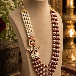 May include: A multi-strand necklace featuring alternating rows of white pearls and deep red beads. The piece includes a gold-toned metal centerpiece depicting two figures playing flutes beneath a small red and gold architectural structure.