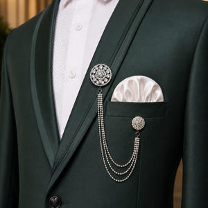 Peut inclure: Un blazer vert foncé avec un revers châle en satin, porté sur une chemise blanche texturée à boutons. Une broche argentée avec trois chaînes en cristal drapées est épinglée au revers, et une pochette blanche est dans la poche poitrine.