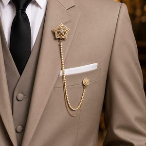 Peut inclure: Une veste de costume beige avec une chemise blanche et une cravate noire, ornée d'une broche dorée en forme d'étoile reliée par deux chaînes fines à une épingle ronde dorée fixée sur la poche poitrine contenant une pochette blanche.