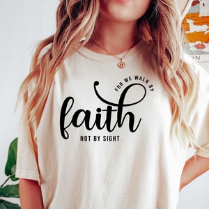 Puede incluir: Una camiseta de color crema con un gr&aacute;fico negro que dice "For we walk by faith not by sight".