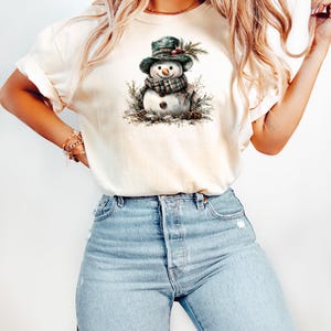 Puede incluir: Camiseta color crema con una ilustraci&oacute;n de acuarela de un mu&ntilde;eco de nieve. El mu&ntilde;eco de nieve lleva una bufanda y un sombrero a cuadros verdes, rodeado de agujas de pino y pi&ntilde;as. La camiseta se combina con vaqueros de mezclilla azul claro.