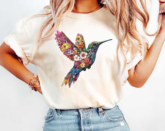 May include: A white t-shirt with a colorful hummingbird graphic. The hummingbird is made up of flowers and leaves.