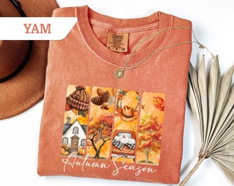 May include: A coral-colored Comfort Colors t-shirt with a graphic of autumn scenes, including a house, trees, and a truck carrying pumpkins. The words "Autumn Season" are written in cursive. A gold necklace is draped across the shirt.