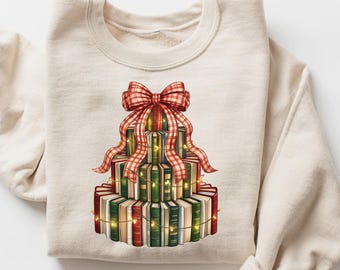 May include: A cream-colored sweatshirt featuring a graphic of a tiered stack of books resembling a cake. The books are in shades of green, red, and brown, adorned with a red plaid bow and string lights. A festive design for book lovers.