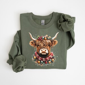 Pu&ograve; includere: Felpa verde oliva con un'illustrazione ad acquerello di una mucca delle Highlands decorata per le feste. La mucca ha una ghirlanda festosa e una sciarpa a quadri. Il disegno &egrave; centrato sulla parte anteriore del top a maniche lunghe.