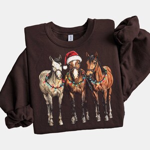 May include: A dark brown sweatshirt featuring a graphic of three horses adorned with Christmas lights. The center horse wears a Santa hat. The horses are in a row, with a white, brown, and chestnut horse.