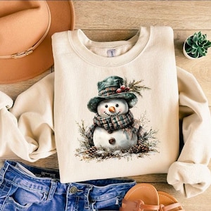 Puede incluir: Sudadera color crema con una ilustraci&oacute;n de acuarela de un mu&ntilde;eco de nieve con bufanda y sombrero a cuadros. El mu&ntilde;eco de nieve est&aacute; rodeado de agujas de pino y pi&ntilde;as. La sudadera se combina con vaqueros azules y un sombrero marr&oacute;n.