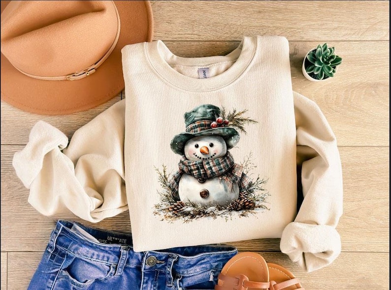 Puede incluir: Sudadera color crema con una ilustraci&oacute;n de acuarela de un mu&ntilde;eco de nieve con bufanda y sombrero a cuadros. El mu&ntilde;eco de nieve est&aacute; rodeado de agujas de pino y pi&ntilde;as. La sudadera se combina con vaqueros azules y un sombrero marr&oacute;n.
