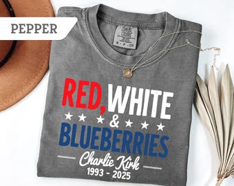 May include: A gray t-shirt with the words "RED, WHITE & BLUEBERRIES" in red, white, and blue. The text "Charlie Kirk 1993-2025" is below. A silver necklace and a brown hat are also visible.