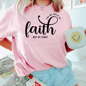 Puede incluir: Camiseta rosa con el texto "For we walk by faith not by sight" en letras negras.