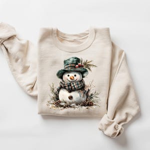 Puede incluir: Sudadera de color crema con una ilustraci&oacute;n en acuarela de un mu&ntilde;eco de nieve. El mu&ntilde;eco de nieve lleva un sombrero y una bufanda a cuadros verdes, con una nariz de zanahoria y ojos de bot&oacute;n. El dise&ntilde;o incluye pi&ntilde;as y vegetaci&oacute;n, evocando un tema de vacaciones de invierno.
