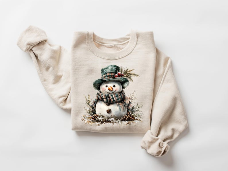 Puede incluir: Sudadera de color crema con una ilustraci&oacute;n en acuarela de un mu&ntilde;eco de nieve. El mu&ntilde;eco de nieve lleva un sombrero y una bufanda a cuadros verdes, con una nariz de zanahoria y ojos de bot&oacute;n. El dise&ntilde;o incluye pi&ntilde;as y vegetaci&oacute;n, evocando un tema de vacaciones de invierno.