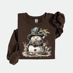 Puede incluir: Una sudadera marr&oacute;n de manga larga con una ilustraci&oacute;n de acuarela de un mu&ntilde;eco de nieve. El mu&ntilde;eco de nieve lleva un sombrero y una bufanda a cuadros verdes, con una nariz de zanahoria y ojos de bot&oacute;n. Pi&ntilde;as y follaje rodean al mu&ntilde;eco de nieve.