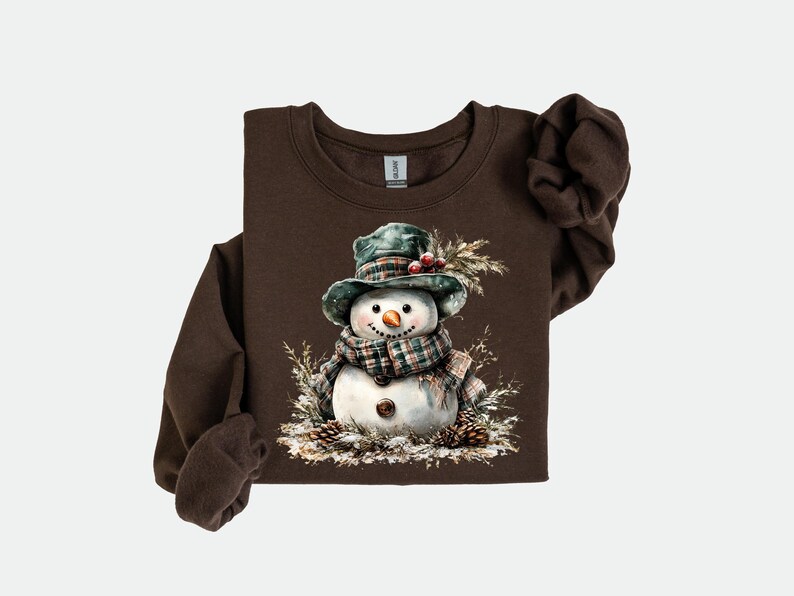Puede incluir: Una sudadera marr&oacute;n de manga larga con una ilustraci&oacute;n de acuarela de un mu&ntilde;eco de nieve. El mu&ntilde;eco de nieve lleva un sombrero y una bufanda a cuadros verdes, con una nariz de zanahoria y ojos de bot&oacute;n. Pi&ntilde;as y follaje rodean al mu&ntilde;eco de nieve.