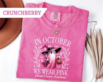 May include: A pink t-shirt with the text "IN OCTOBER WE WEAR PINK Breast Cancer Awareness". The shirt features a ghost wearing a witch hat and holding a pumpkin. A gold necklace is draped across the shirt.