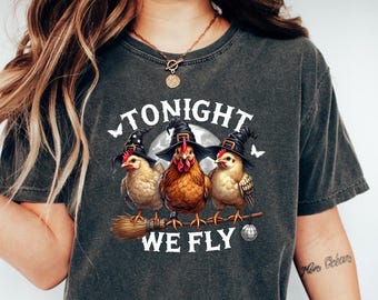 May include: Dark gray t-shirt featuring three chickens wearing witch hats, perched on a broomstick, with the text "TONIGHT WE FLY" above and below them. A full moon and butterflies are in the background. The shirt is a comfortable, casual style.